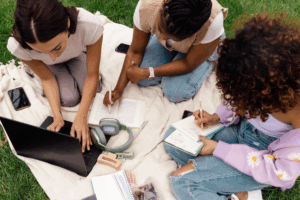 Three college students studying together on a blanket outdoors with a laptop, books, and notebooks — ideal for student budgeting, college financial planning, or credit-building tips.