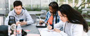 Three college students studying outdoors with books, tablets, and water bottles on a campus table — ideal image for financial education, budgeting, or student credit-building topics.