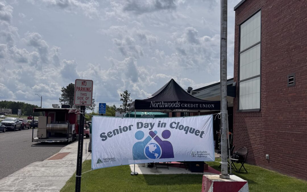 Outdoor scene of a community event with a tall flagpole and waving American flag beside a brick building and a banner that reads 'Senior Day in Cloquet' under a black canopy tent.
