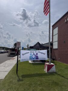 Outdoor scene of a community event with a tall flagpole and waving American flag beside a brick building and a banner that reads 'Senior Day in Cloquet' under a black canopy tent.